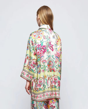 A person with long blonde hair stands against a plain background, wearing the MIRTO 1956 - FLUID BLOUSE MULTICOLOR FLORAL by MIRTO 1956. The uplifting blouse features a vibrant, floral pattern in pink, green, yellow, and blue. The 3/4 sleeves and V neckline enhance the intricate designs, creating a bright and stylish look.