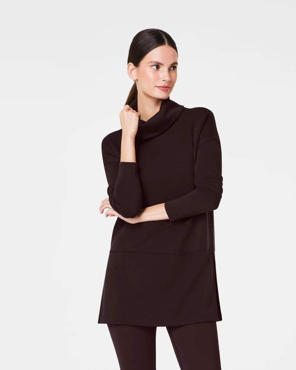 Dressed in the luxurious comfort of a SPANX AireEssentials Turtleneck Tunic, characterized by its black hue and lightweight fabric, a person stands with their back to the camera. Their elegantly tied up hair complements the matching black pants against a plain white backdrop.