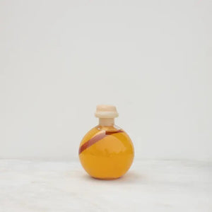 A round glass bottle filled with orange liquid, sealed with a wooden cork, and tied with a string. A white label with small text is attached to the bottle, potentially indicating that it is "STONE HOLLOW FARMSTEAD - INFUSED VINEGAR HOT PEPPER" by STONE HOLLOW FARMSTEAD. The bottle is set against a plain, light gray background.