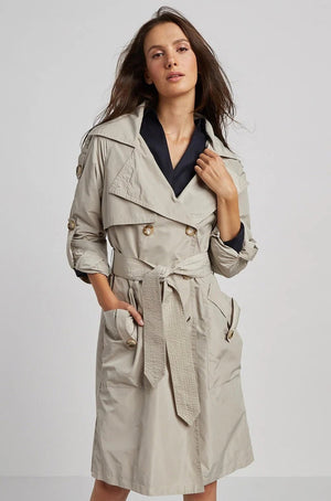 A woman with long brown hair poses in the ADROITE ATELIER - RAPHAEL TRENCH COAT by ADROIT ATELIER. This light beige trench coat highlights rolled-up sleeve details, a belted waist, and a flap on the back. She stands against a plain gray background, glancing over her shoulder.