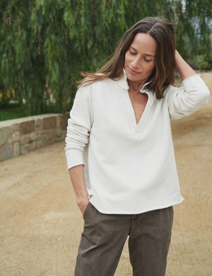 A person with long hair stands outdoors, wearing a vintage white Patrick Popover Henley in triple fleece from Frank & Eileen. Their sleeves are casually rolled up and their arms crossed, while a path and greenery can be seen in the blurred background.