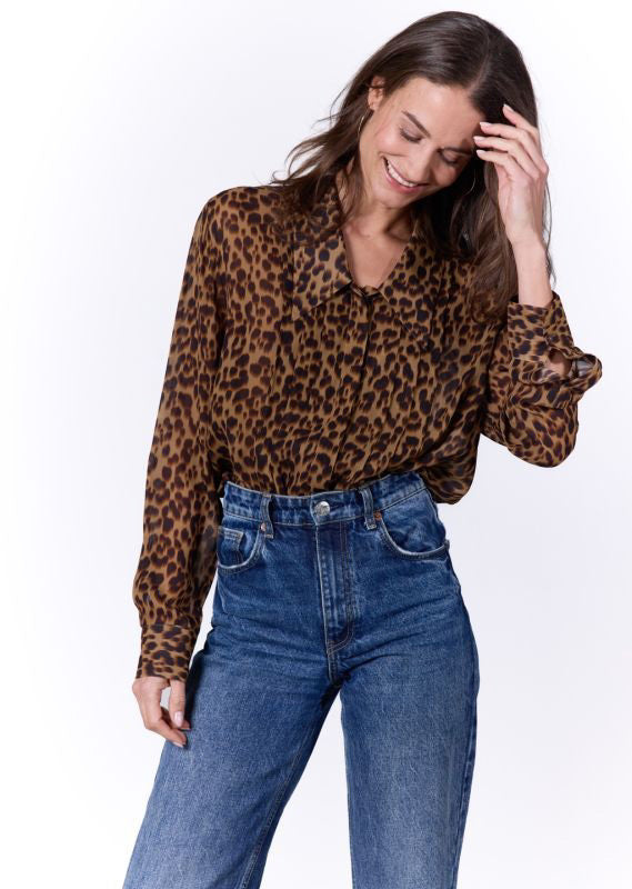 A woman with long brown hair smiles and looks down while wearing the 0039 ITALY - JOLA BLOUSE, a viscose chiffon leopard print blouse, paired with high-waisted blue jeans. She has her right hand raised to her hair and stands in front of a plain white background.