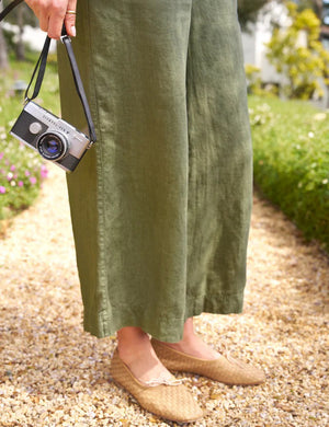 A woman wearing an olive green Tuscany Getaway Set, featuring the FRANK & EILEEN Maisie Wide Leg Linen Pull-On Pant, and a wide-brimmed straw hat strolls along a gravel path in a lush garden. With her hands in her pockets and a relaxed expression, she looks to the side, enjoying the serene ambiance.