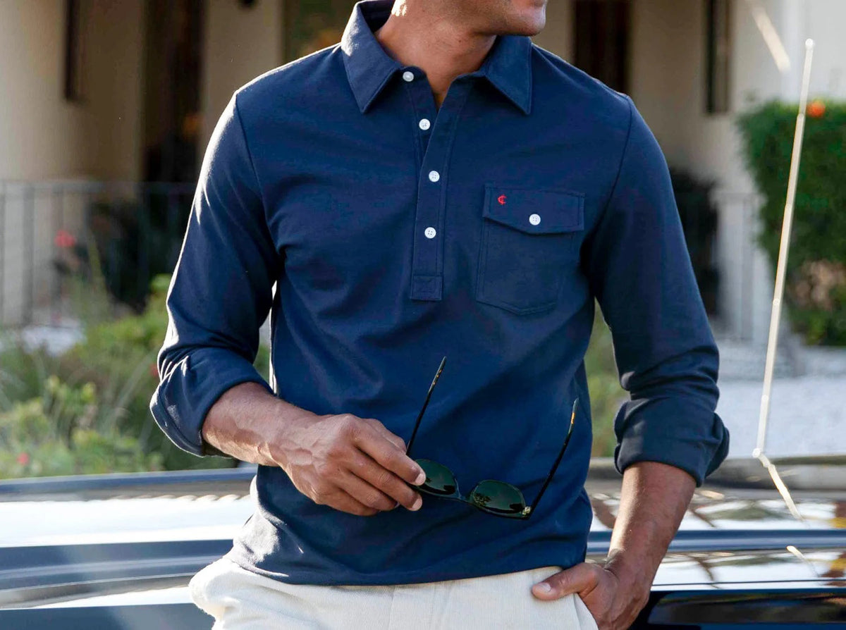 A man wears the CRIQUET Long Sleeve Players Polo Shirt in Peacoat, made from soft Peruvian Pima cotton with a buttoned collar and chest pocket. He stands with one hand in his jeans pocket against a white background.