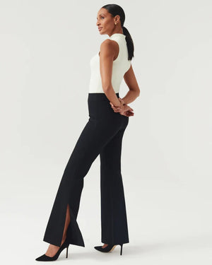 A woman stands posed against a light background, wearing the SPANX - PERFECT PANT SPLIT HEM, featuring black high-waisted pants crafted from smoothing premium ponte fabric with slit flares. Paired with a white sleeveless top and black high heels, her hair is pulled back as she looks to the side, hands clasped gracefully behind her back.