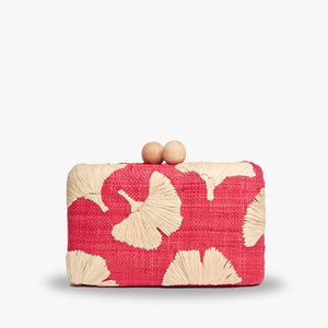 The KAYU AMI CLUTCH is a small pink raffia clutch with tan floral patterns, a beige interior, an optional gold chain strap, and a clasp featuring two wooden beads. The hand-embroidered charm is highlighted by the "Kayu" label inside.