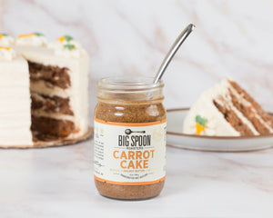 A 13-ounce (369 grams) jar of BIG SPOON ROASTERS - CARROT CAKE nut butter with sea salt. The jar features a white label adorned with orange accents and the BIG SPOON ROASTERS logo at the top. This vegan, handcrafted spread, rich in heart-healthy fats, is sealed with a gold lid.