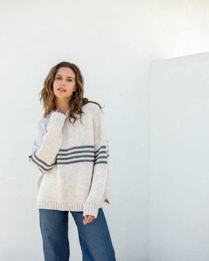 A person with long, wavy brown hair is wearing the MERSEA - SEACOAST STRIPE SWEATER, a loose-fitting, cozy blue knit sweater with white stripes on the sleeves, paired with black pants. They are standing against a plain, light-colored background and facing left, with their hands near their face.