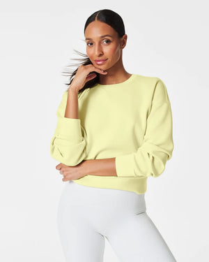 A woman stands confidently against a plain white background, wearing the SPANX Aireessentials Crew Neck Pullover and matching sweatpants made from luxurious comfort spacer fabric. She has her hair pulled back and her left hand is raised slightly. She smiles gently with her lips closed.