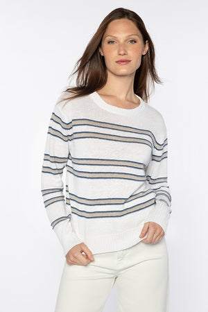 A person with long brown hair stands against a plain white background, wearing a cozy KINROSS CASHMERE - LINEN STRIPE CREW PULLOVER from KINROSS, featuring horizontal blue and gray stripes. They paired it with white pants. Their right hand is in their pocket while their left arm rests by their side.