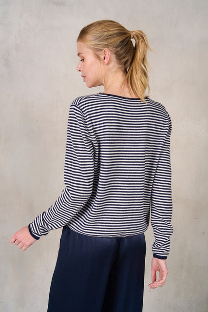 A person with blonde hair in a ponytail is shown from the back, wearing the ROSE & CROWN STRIPE CASHMERE CREWNECK SWEATER and dark blue trousers. The outfit exudes a sophisticated look against the plain, light-colored wall backdrop.