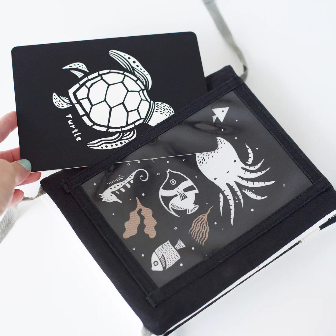 A TUMMY TIME GALLERY soft book from WEE GALLERY, featuring black and white fish and squid illustrations, is partially inserted in a clear front pocket. A brown tag attached by a string reads &quot;tummy time gallery.&quot; This Montessori toy, ideal for sensory development, rests on a plain white surface.