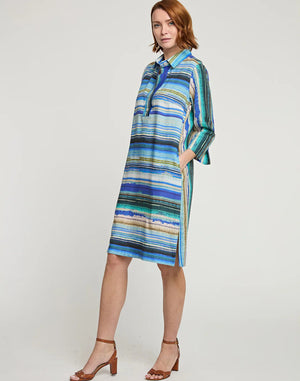 A woman stands against a plain background wearing the HINSON WU - CHARLOTTE 3/4 SLEEVE TENCEL TEXTURED STRIPE DRESS, a knee-length dress with 3/4 sleeves and colorful horizontal stripes in shades of blue, green, and beige. Featuring a shirt collar and Tencel textured stripes, she has one hand in her dress pocket and is wearing brown heeled sandals.