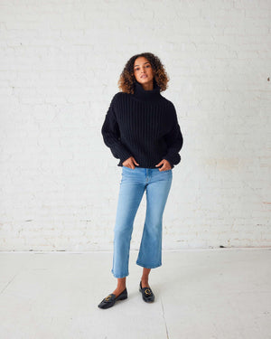 A woman with curly hair stands against a white wall, wearing a chunky mustard-colored MERSEA - BEACHSIDE TURTLENECK and blue jeans. She holds the hem of her turtleneck with one hand and has a relaxed expression.