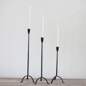 Three tall, lit white candles sit in varying height HAND FORGED CAST IRON FOOTED TAPER HOLDERS by CREATIVE COOP on a wooden surface against a plain light background.