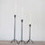 Three tall, lit white candles sit in varying height HAND FORGED CAST IRON FOOTED TAPER HOLDERS by CREATIVE COOP on a wooden surface against a plain light background.