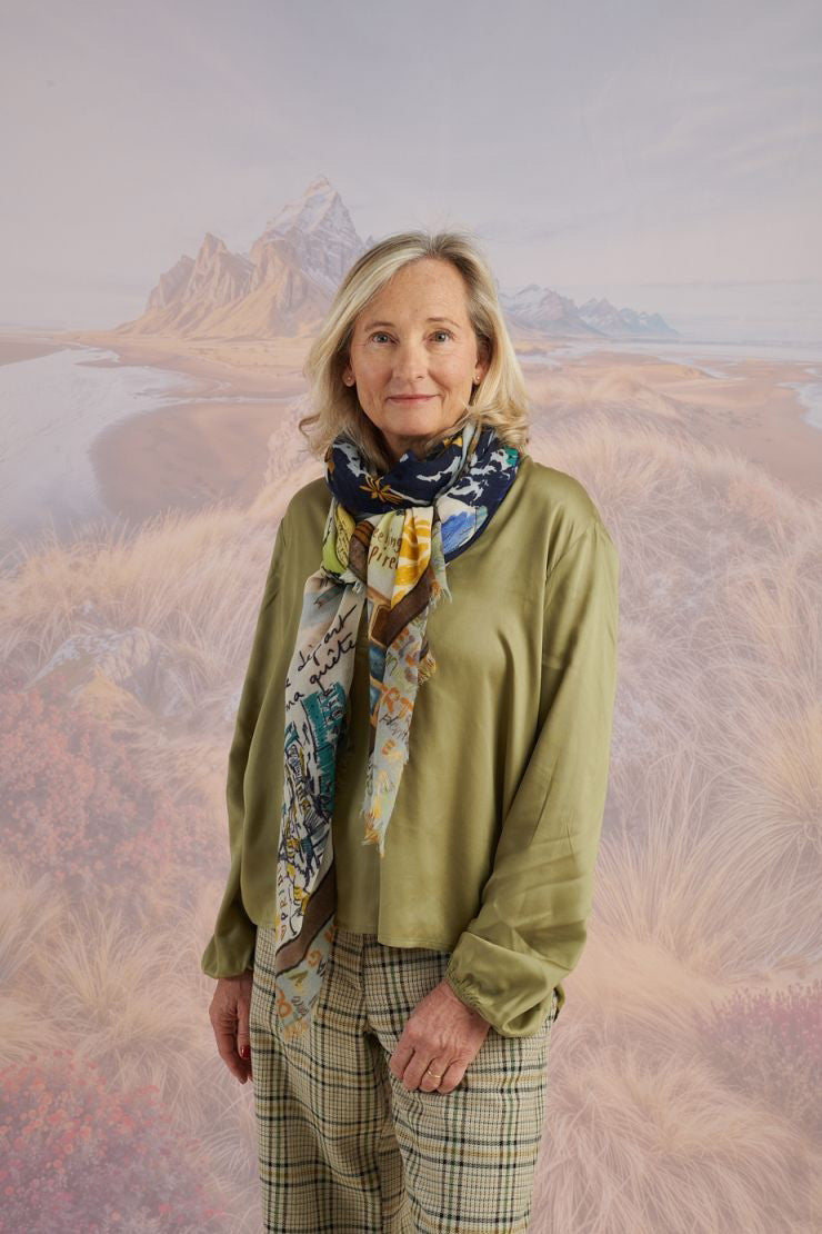 A woman with wavy blonde hair smiles while looking down. She wears a loose, light lavender blouse, dark pants, and the STORIATIPIC EUGENIE WOOL SCARF. Behind her is a scenic landscape of mountains, grass, and water.