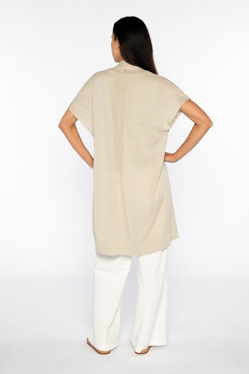 A woman with long dark hair, hands on hips, wears the KINROSS CASHMERE - LONG SHORT SLEEVE LINEN CARDIGAN in beige over white pants. Her back faces the camera against a plain white backdrop.