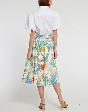 A person is wearing a white button-up shirt with short, puffy sleeves, and a high-waisted GLORIA SKIRT by HINSON WU featuring a vibrant Rainforest Print in various colors. They are also wearing tan, open-toe sandals with a low heel. The background is plain white.