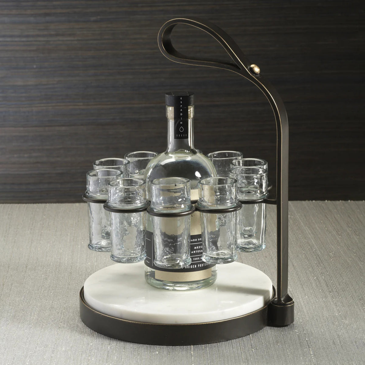 The ZODAX TEQUILERO 10 Shot Tequila Set features a circular white marble cutting board with an iron handle and metal frame, holding ten empty shot glasses arranged in a ring.