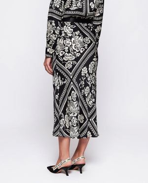 The MIRTO 1956 - BLACK AND WHITE PRINT MIDI SKIRT by MIRTO 1956 features a black A-line silhouette with an intricate white floral squares patchwork design set within diamond shapes. This skirt has a slight flare, providing a flowy appearance, and its symmetrical pattern offers a balanced and flattering chic look.