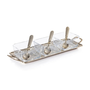 The ROUSSILLON CONDIMENT SET by ZODAX features three clear glass bowls with brass spoons, displayed on a rectangular gold metal tray with handles against a white background.