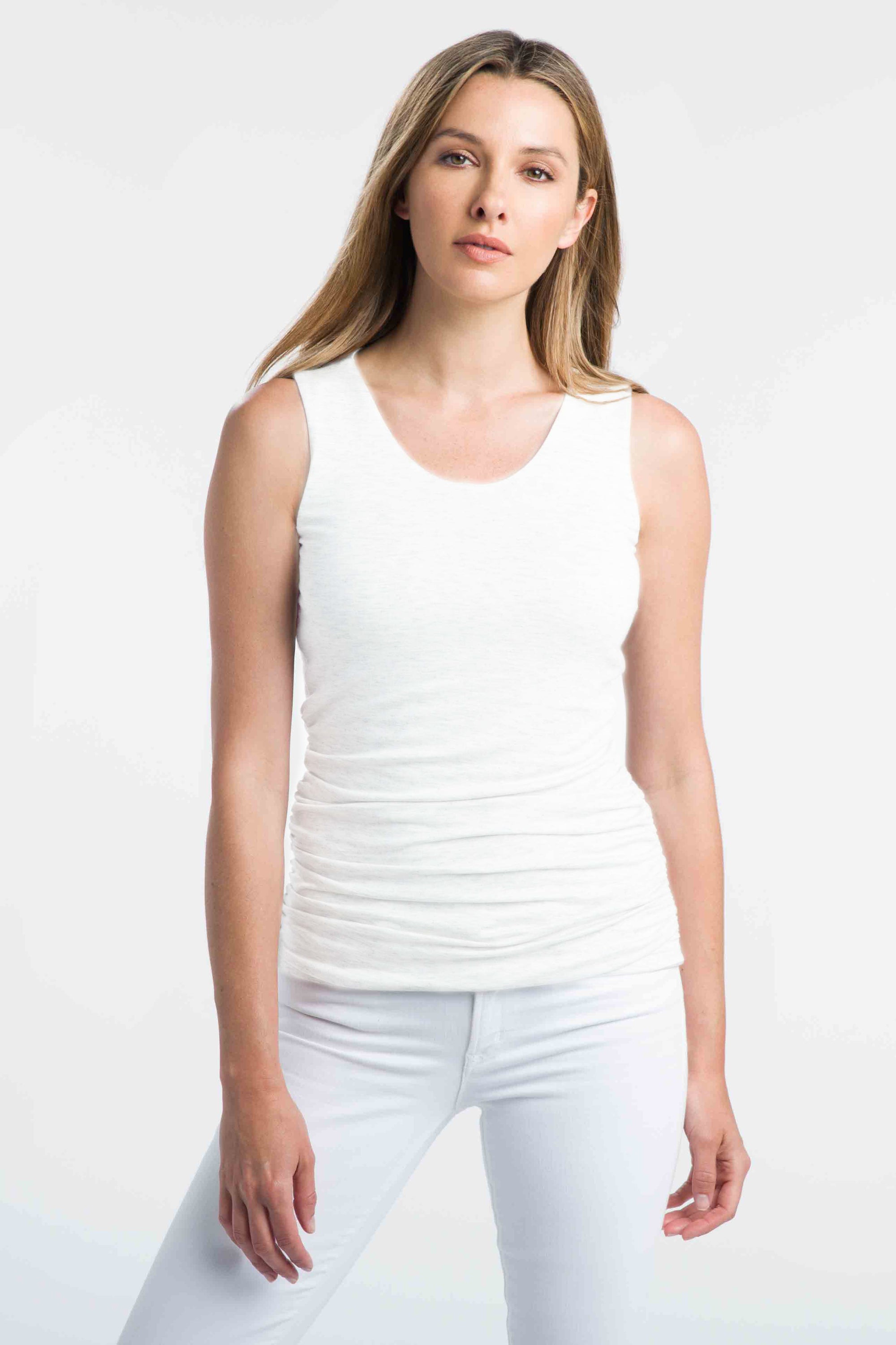 A woman with long, blonde hair stands against a plain white background. She is wearing the KINROSS CASHMERE - COTTON RUCHED TANK IN WHITE and matching white pants. Her look exudes an effortless elegance, reminiscent of the timeless pieces from KINROSS. She has a neutral expression and her arms are relaxed at her sides.