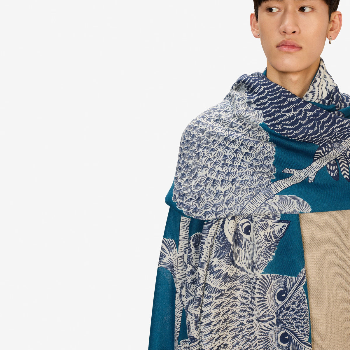A blonde woman in a black coat stands against a white background, wearing the INOUI EDITIONS &quot;Owls on a Branch&quot; wool scarf in blue by INOUI, draped over her neck and one shoulder with intricate white owl designs.