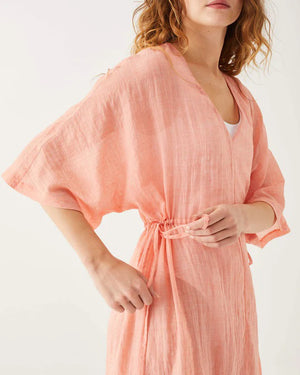 A woman in a light pink MER SEA - BREEZY KAFTAN DRESS, featuring slits and a cinched waist, walks barefoot. She has long, wavy hair and is smiling, facing slightly to the right. The background is plain white.