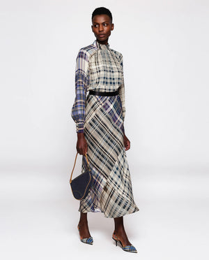 A person is modeling an uncomplicated chic outfit that includes the MIRTO 1956 - BLUE PLAID MIDI SKIRT paired with a long-sleeved, high-necked blouse, pointed heels, and holding a small, dark handbag. The skirt features a cinched waist and long design, and the person has short hair.