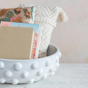 A white textured terra cotta bowl with raised dots from Creative Coop contains a beige book, a blue booklet, and a pink booklet. It stands on a distressed white wooden surface, enhancing the rustic charm. Behind the bowl are two decorative pillows with different patterns: one featuring colorful floral embroidery and the other with a cream-colored knit design.