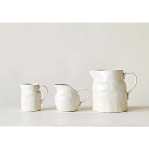 The STONEWARE VINTAGE PITCHER by CREATIVE COOP is a white ceramic piece with a smooth, wavy texture and curved handle, ideal for serving drinks or displaying flowers. It blends minimalist design with vintage-inspired charm.