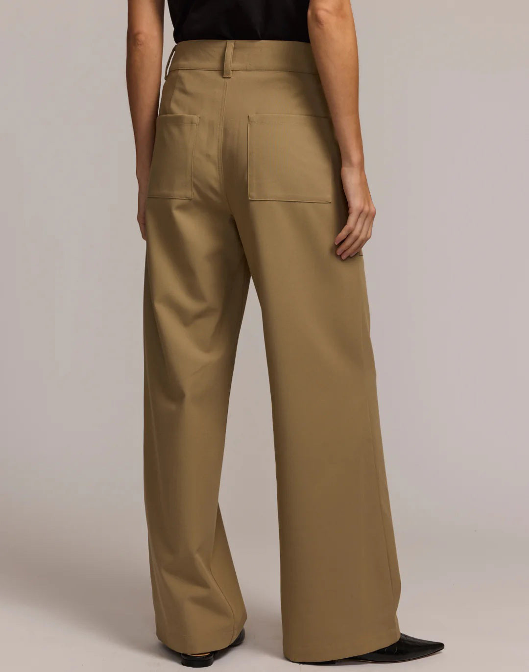 A person wears the HINSON WU - KELLY TRAVELER&#39;S TWILL PANT—wide-leg khaki trousers with two front pockets and double button closure. Hands are in the pockets, and shoes are partially visible.