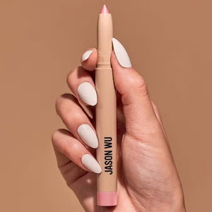 Close-up of a hand with white painted nails holding a sparkle pearl colored eyeshadow pencil with a pink tip, branded with "JASON WU BEAUTY" in black text against a solid neutral beige background.