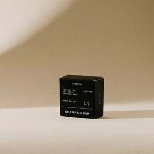 A small, black rectangular box of sulfate-free fortifying tea tree shampoo bar stands against a neutral beige background. The packaging reads "PRESTON - SHAMPOO BAR, Fortifying Tea Tree Leather Shampoo Bar, Made in USA, 3 oz, 85.6 g," highlighting its plastic-free and plant-based cleansers.