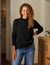 A person with long curly hair wearing a black FRANK & EILEEN Monterey Rolled Funnel Neck Sweater and blue jeans stands by a wooden dresser with a lit candle on it. The room has a cozy, muted tone, accentuated by the textured wall and gentle lighting.