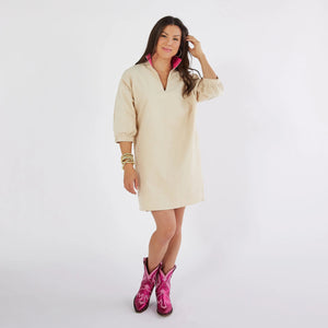 A woman stands against a plain white background wearing the CARYN LAWN - BETSY COLLAR CORDUROY DRESS, which features a relaxed fit in beige with a pink collar. She pairs it with vibrant pink cowboy boots and accessorizes her ensemble with bracelets on her left wrist. Her long brown hair cascades down as she smiles gently at the camera in this one-size-fits-all outfit by CARYN LAWN.