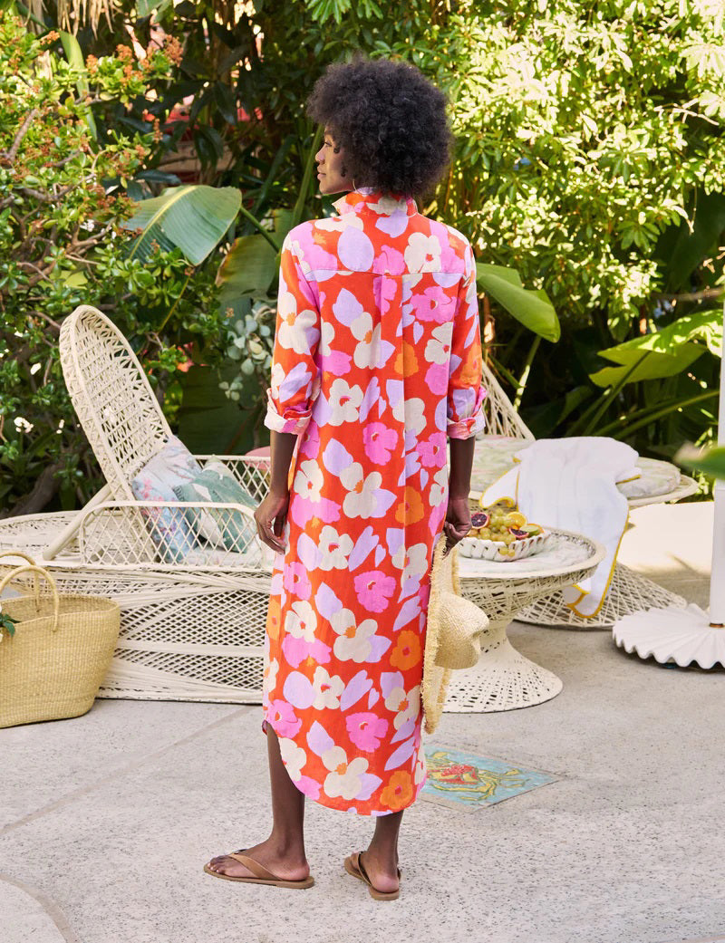 A person with curly hair stands outdoors, wearing the FRANK &amp; EILEEN Rory Maxi Shirtdress in Classic Italian Linen—Orange and Pink Floral—embodying California chic amid lush greenery and white outdoor furniture, holding a woven straw bag.