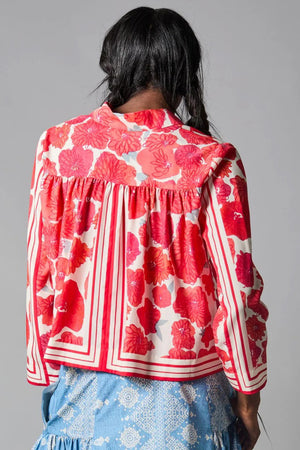 A person with dark braided hair wears the HOPE FOR FLOWERS - SCARF PRINT SHIRT, showcasing red and white floral patterns with striped accents. This loose-fitting blouse, crafted from sustainable REFIBRA fabric, is paired with a blue skirt featuring lace details against a plain gray background.