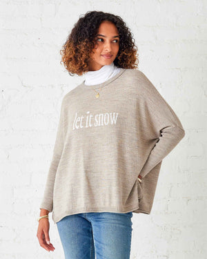 A person with curly hair is wearing the MERSEA - LET IT SNOW CATALINA CREWNECK SWEATER in beige over a white shirt and blue jeans, standing sideways against a white brick wall. They are smiling and looking at the camera.