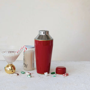 Flat lay of festive cocktail ingredients on a white background, highlighting the 22 oz. enameled stainless steel cocktail shaker by Creative Coop, a gold-rimmed glass with a pink drink, disco balls, colorful sweets, macarons, red spoon, and striped straw. A small cactus and napkin with a drink umbrella add flair.