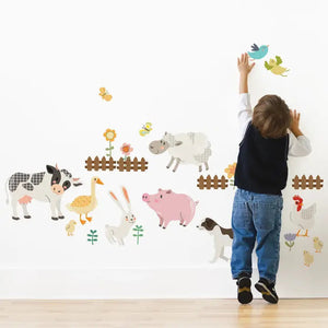 A young child reaches up towards a blue bird decal on a white wall decorated with SIMPLE SHAPES - FARM ANIMALS REMOVABLE WALL DECALS. The wall showcases decals of a cow, sheep, duck, rabbit, pig, dog, and chicken, along with flowers and butterflies from the SIMPLE SHAPES brand, creating a playful farm scene.