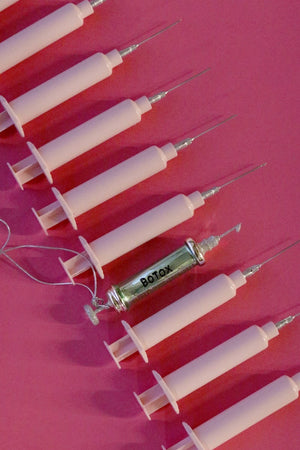 An arrangement of glass syringes on a pink background features a central syringe labeled "Wrinkle Free Ornament." These precision-crafted syringes, part of the CODY FOSTER brand, are aligned in a row with the primary one positioned horizontally among the others standing vertically.