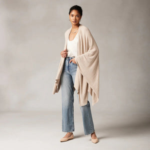 A person wearing the KASHWERE - TRAVEL PONCHO, a beige, draped poncho with a cowl neck and wide sleeves, poses with their arms raised and hands touching the back of their head. They have a neutral expression and are standing against a plain background.