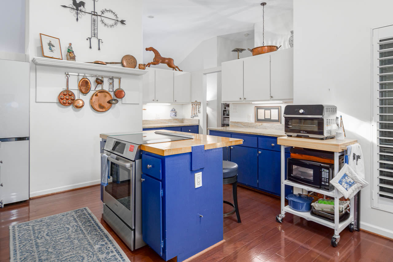 Bright kitchen with blue lower cabinets, stainless steel stove, butcher block countertops, hanging copper pans, white upper cabinets, a rolling cart with a microwave and toaster oven, and wooden floors. Decor items sit on shelves.