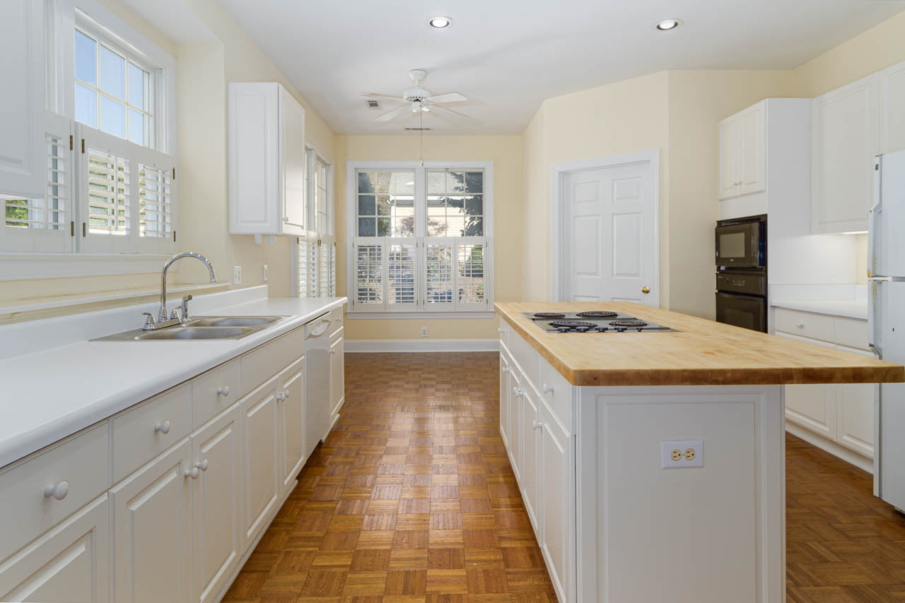 Bright kitchen with white cabinets, a wooden island countertop with a stove, built-in oven, double sink, large windows with shutters, and parquet wood flooring.