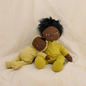 An adorable DOZY DINKUM DOLL by OLLI ELLA USA, featuring soft brown fabric skin, closed embroidered eyes, and short black hair, is dressed in a charming yellow onesie adorned with a rainbow design on the chest. Perfect as a first toy, the doll is lying on a white, quilted surface.