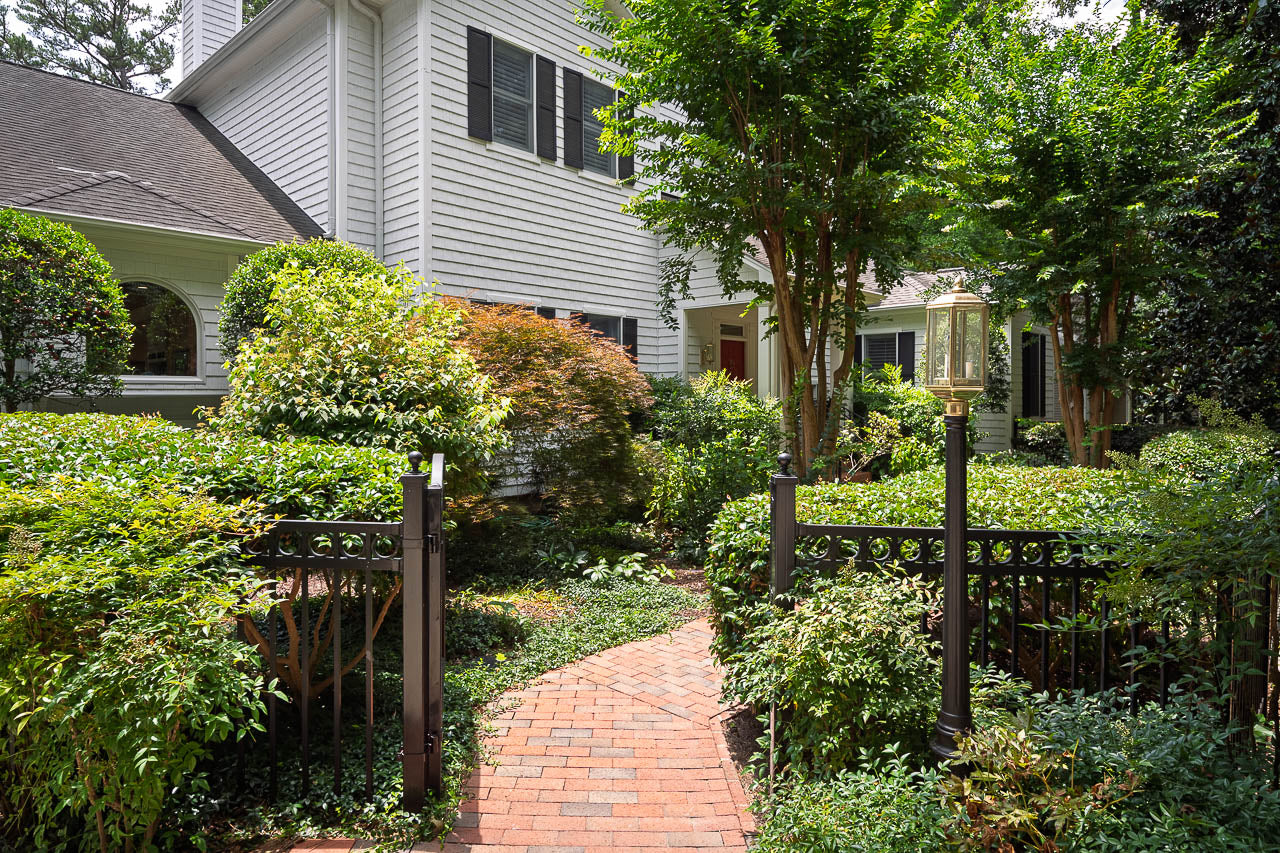 A brick walkway leads through a black metal gate and lush green garden to the entrance of a white two-story house with black shutters and a porch, surrounded by trees and shrubs.