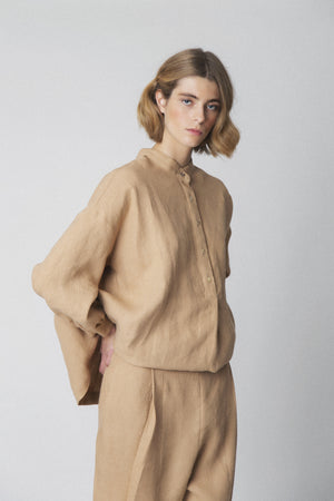 A person with short, wavy blonde hair wears the LOESS - FLORENCE LINEN BLOUSE in beige. Featuring gathered fabric detail on the back, it stands out against a neutral light gray background, showcasing timeless style and sustainable fashion by LOESS.