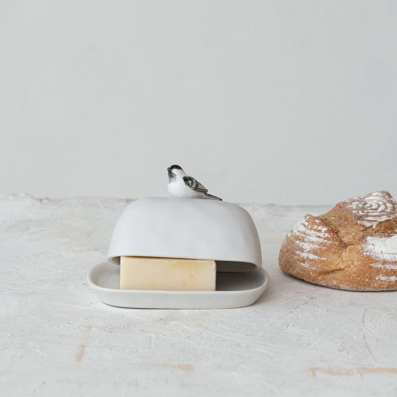 The STONEWARE BUTTER DISH WITH BIRD HANDLE by CREATIVE COOP measures 7"L x 4-3/4"W x 4"H and features a charming bird-shaped handle on the lid. When lifted, it reveals a block of butter inside. The dish sits gracefully on a light-colored surface, with an artisan loaf of bread partially visible in the background.